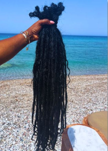 A bundle coolie locs with curly raw Indian hair extensions held up by a hand against a sunny beach backdrop with turquoise water and pebbled sand.