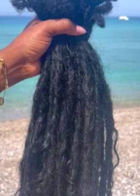 A bundle coolie locs with curly raw Indian hair extensions held up by a hand against a sunny beach backdrop with turquoise water and pebbled sand.
